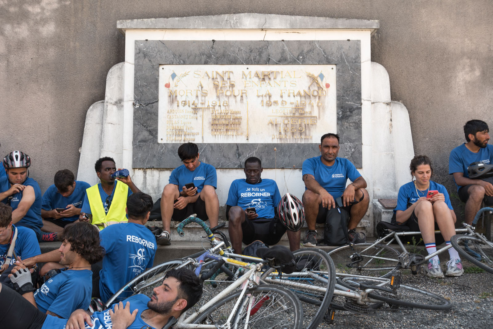 Refugees and volunteers rest in the shade in front of a war memorial at Saint-Martial in the Nouvelle-Aquitaine region of France, during the Reborn Trip bicycle crossing of France on August 8, 2024. Des refugies et des benevoles se reposent a l ombre devant un monument aux morts a Saint-Martial en Nouvelle-Aquitaine pendant la traversee de la France a velo de Reborn Trip le 8 aout 2024.