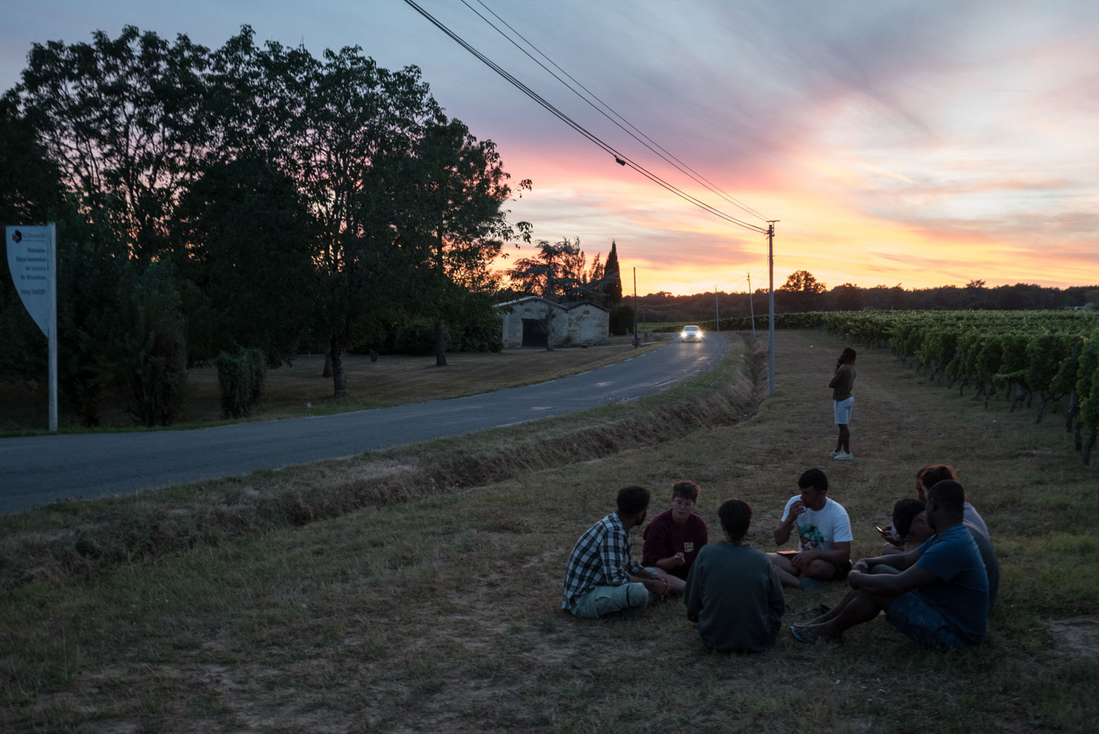 A group of volunteers and refugees share a moment at sunset as they sit on the ground in a vineyard beside a road next to the Blasimon campsite, where they are staying for the night, during the Reborn Trip bicycle tour of France in the Nouvelle-Aquitaine region on August 8, 2024.Un groupe de benevoles et de refugies partagent un moment au coucher du soleil assis par terre dans un champs de vigne au bord d'une route a cote du camping de Blasimon, ou ils logents pour la nuit, pendant la traversee de la France a velo de Reborn Trip en Nouvelle-Aquitaine le 8 aout 2024.